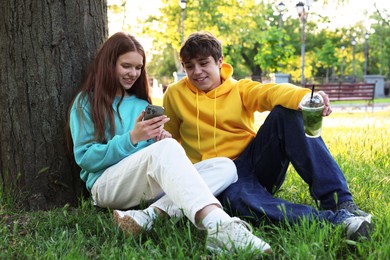 Photo of Teenage girl and boy with smartphone and drink on green grass in park