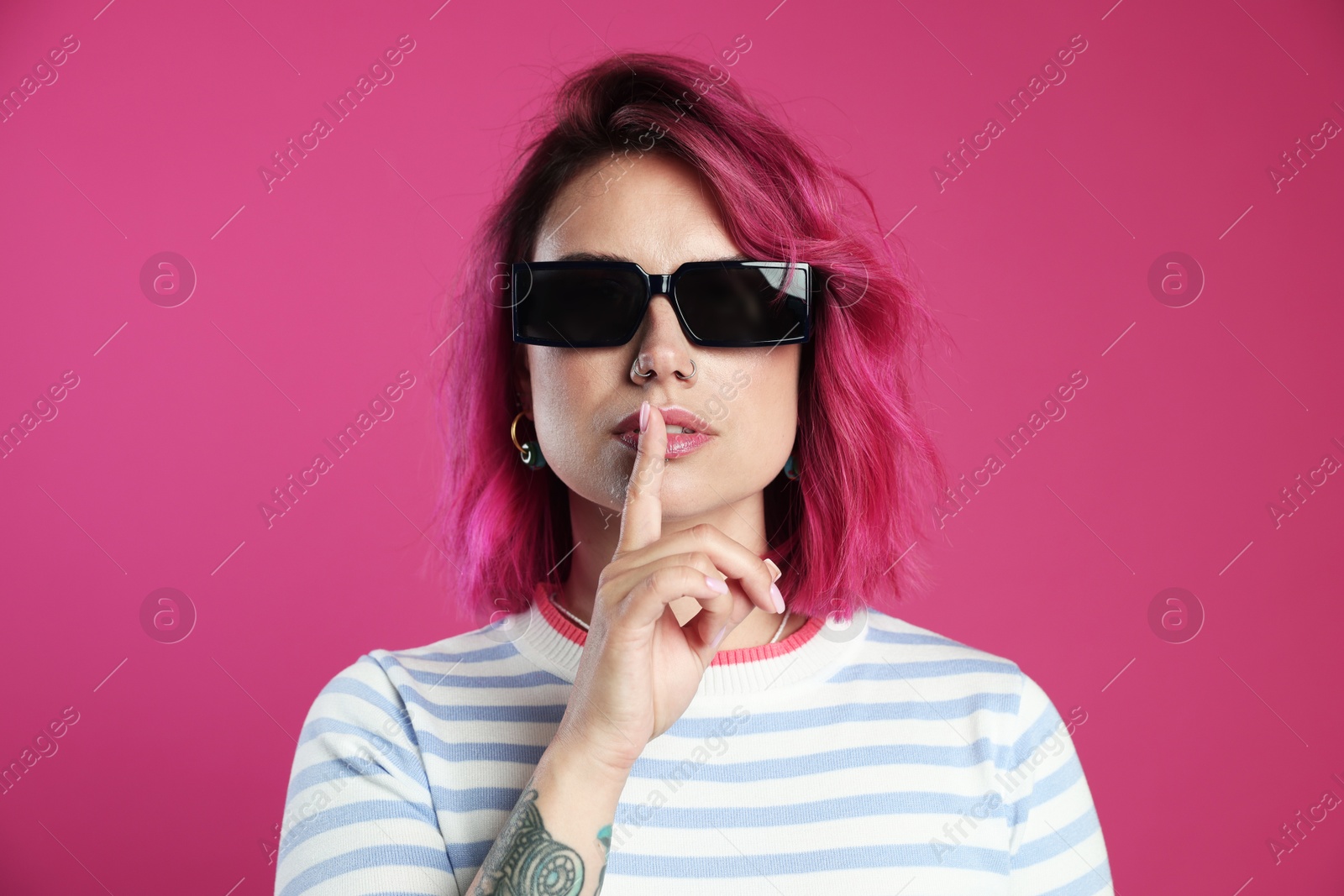 Beautiful woman with bright dyed hair in sunglasses making hush gesture on pink background Photo of Beautiful woman with bright dyed hair in sunglasses making hush gesture on pink background