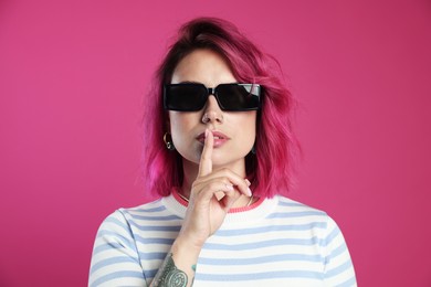 Photo of Beautiful woman with bright dyed hair in sunglasses making hush gesture on pink background