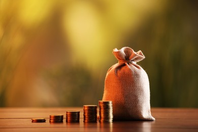 Photo of Burlap bag and stacked coins on wooden table against blurred background