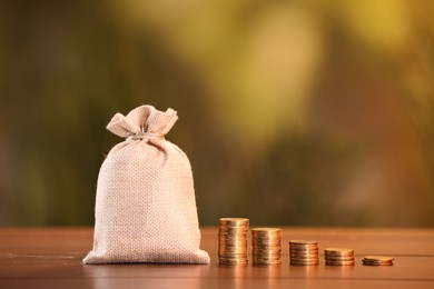 Photo of Burlap bag and stacked coins on wooden table against blurred background