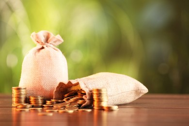 Photo of Burlap bags and stacked coins on wooden table against blurred background, space for text