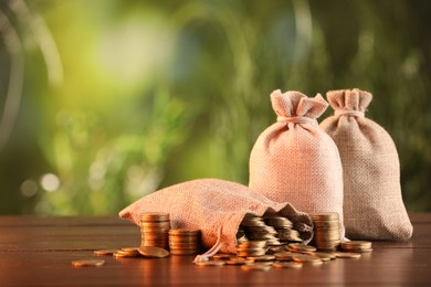 Photo of Burlap bags and stacked coins on wooden table against blurred background, space for text