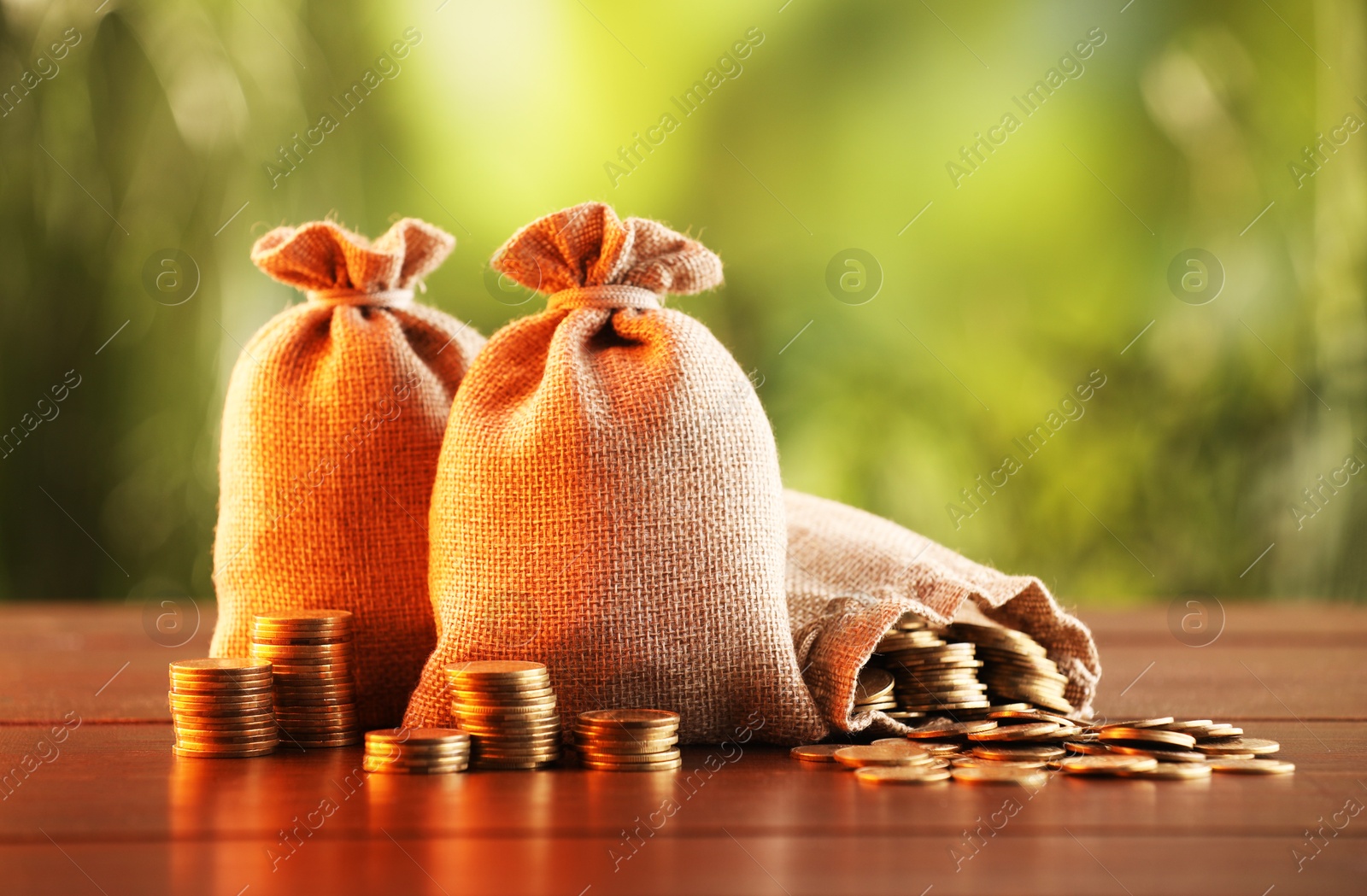 Photo of Burlap bags and stacked coins on wooden table against blurred background