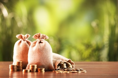 Photo of Burlap bags and stacked coins on wooden table against blurred background, space for text