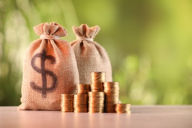 Photo of Burlap bags and stacked coins on table against blurred background, space for text