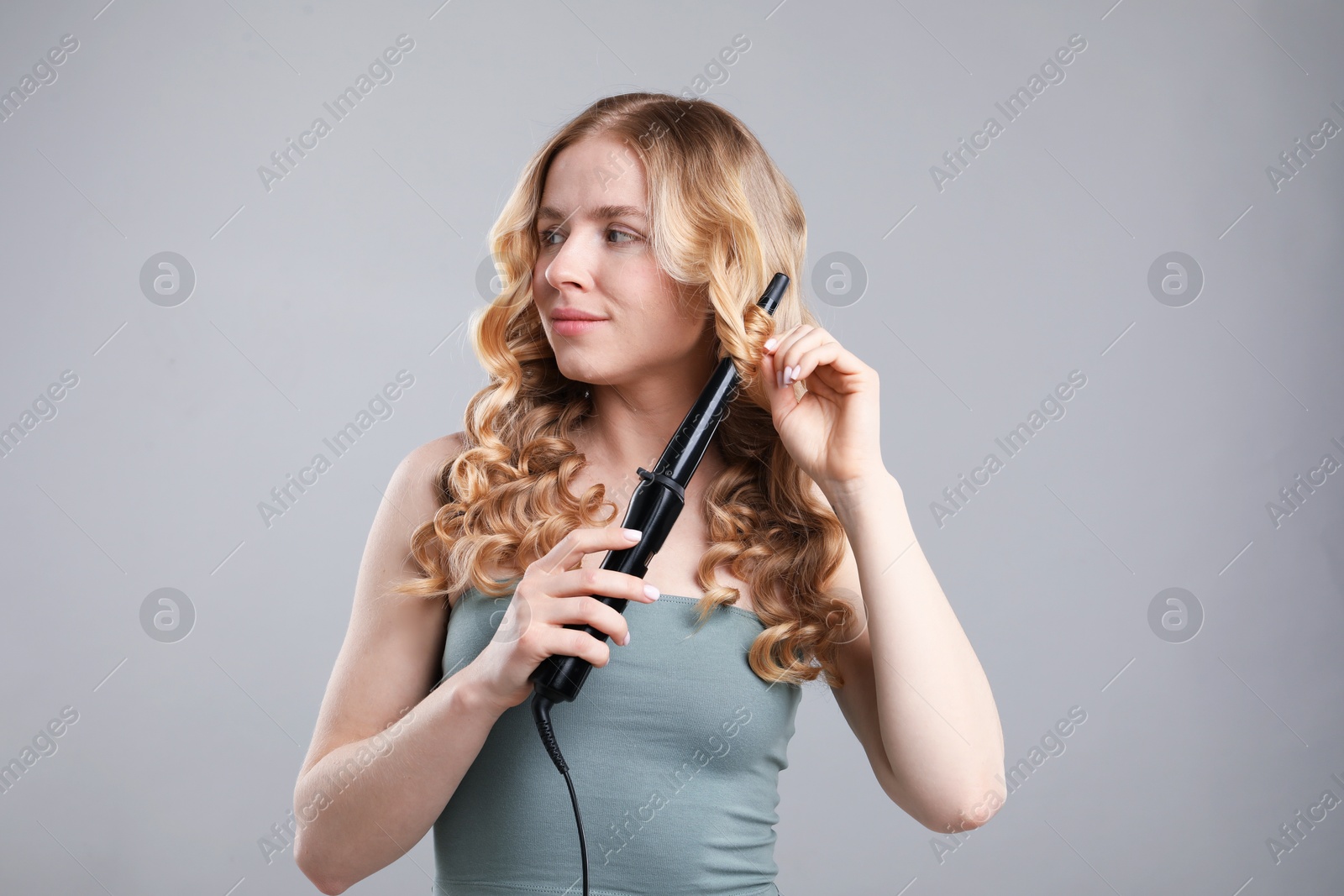 Woman curling her hair on grey background Photo of Woman curling her hair on grey background