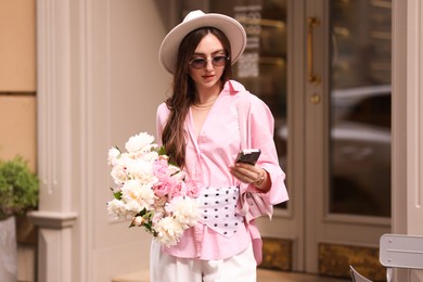 Photo of Stylish woman with bouquet of beautiful peonies and smartphone outdoors