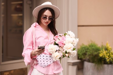 Photo of Stylish woman with bouquet of beautiful peonies and smartphone outdoors