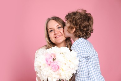Photo of Little boy kissing his mom and giving her beautiful peony flowers on pink background