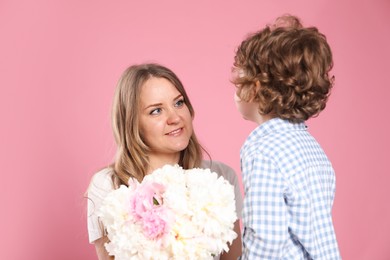 Photo of Little boy giving his mom beautiful peony flowers on pink background