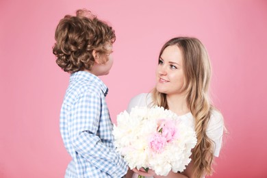 Photo of Little boy giving his mom beautiful peony flowers on pink background