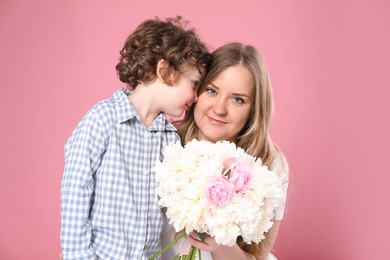 Photo of Little boy giving his mom beautiful peony flowers on pink background