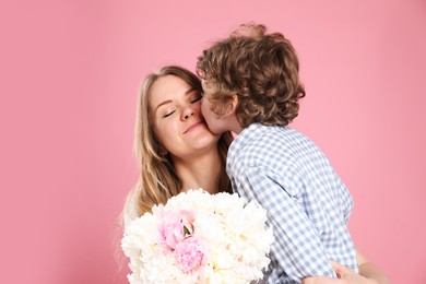 Photo of Little boy kissing his mom and giving her beautiful peony flowers on pink background