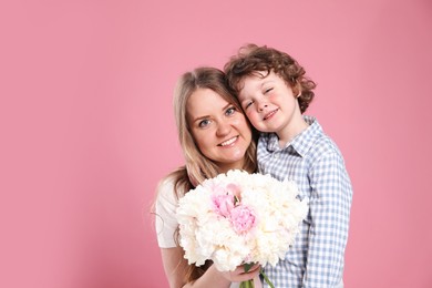 Photo of Little boy giving his mom beautiful peony flowers on pink background. Space for text