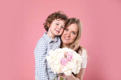 Photo of Little boy giving his mom beautiful peony flowers on pink background