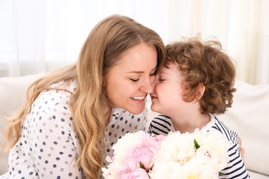 Photo of Little boy giving his mom beautiful peony flowers on sofa at home