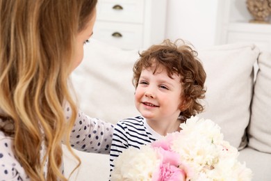 Photo of Little boy giving his mom beautiful peony flowers on sofa at home