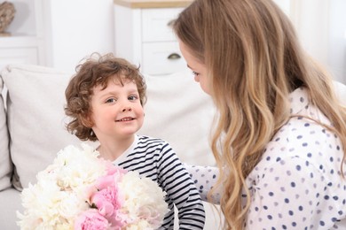 Photo of Little boy giving his mom beautiful peony flowers on sofa at home