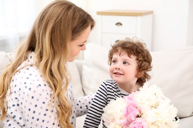 Photo of Little boy giving his mom beautiful peony flowers on sofa at home