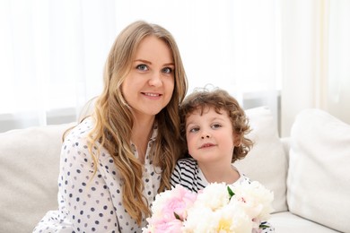 Photo of Little boy giving his mom beautiful peony flowers on sofa at home