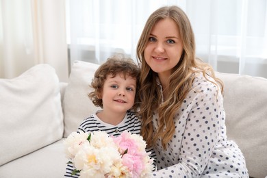 Photo of Little boy giving his mom beautiful peony flowers on sofa at home