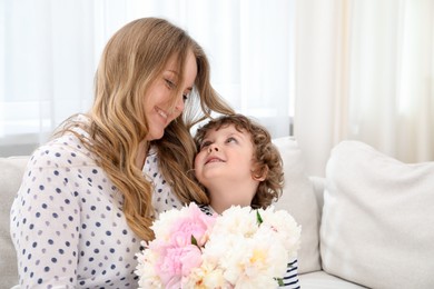 Photo of Little boy giving his mom beautiful peony flowers on sofa at home. Space for text