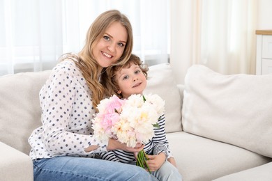 Photo of Little boy giving his mom beautiful peony flowers on sofa at home. Space for text