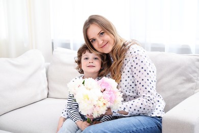 Photo of Little boy giving his mom beautiful peony flowers on sofa at home. Space for text
