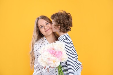 Photo of Little boy kissing his mom and giving her beautiful peony flowers on yellow background