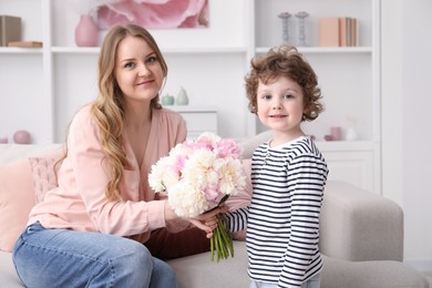 Photo of Little boy giving his mom beautiful peony flowers on sofa at home