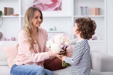 Photo of Little boy giving his mom beautiful peony flowers on sofa at home