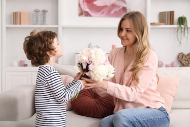 Photo of Little boy giving his mom beautiful peony flowers on sofa at home