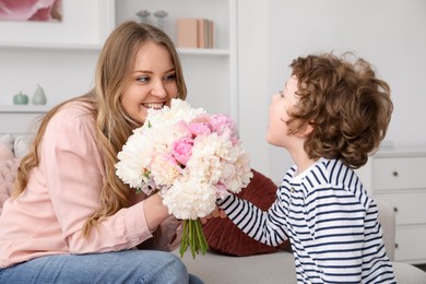 Photo of Little boy giving his mom beautiful peony flowers on sofa at home