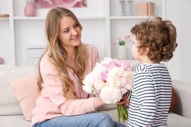 Photo of Little boy giving his mom beautiful peony flowers on sofa at home