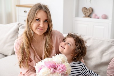 Photo of Little boy giving his mom beautiful peony flowers on sofa at home