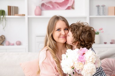 Photo of Little boy kissing his mom and giving her beautiful peony flowers at home. Space for text