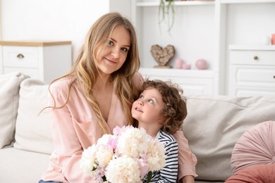 Photo of Little boy giving his mom beautiful peony flowers on sofa at home