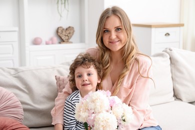Photo of Little boy giving his mom beautiful peony flowers on sofa at home
