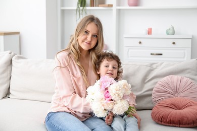 Photo of Little boy giving his mom beautiful peony flowers on sofa at home