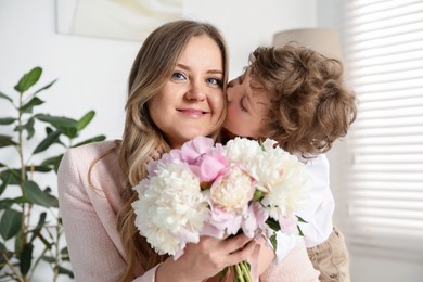 Photo of Little boy kissing his mom and giving her beautiful peony flowers at home