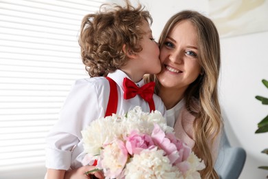 Photo of Little boy kissing his mom and giving her beautiful peony flowers at home