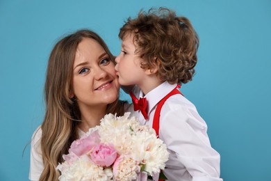 Photo of Little boy kissing his mom and giving her beautiful peony flowers on light blue background