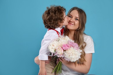 Photo of Little boy kissing his mom and giving her beautiful peony flowers on light blue background