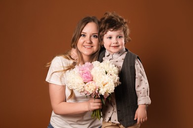 Photo of Little boy giving his mom beautiful peony flowers on brown background