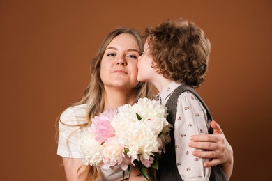 Photo of Little boy kissing his mom and giving her beautiful peony flowers on brown background