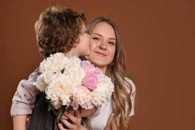 Photo of Little boy kissing his mom and giving her beautiful peony flowers on brown background