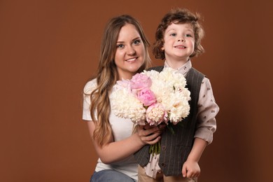 Photo of Little boy giving his mom beautiful peony flowers on brown background