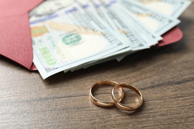 Photo of Gold wedding rings, envelope and dollar banknotes on wooden table, closeup