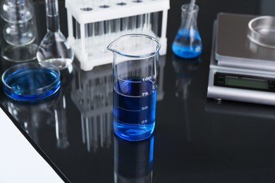 Photo of Beaker with blue liquid, other laboratory glassware and scale on black mirror table, closeup